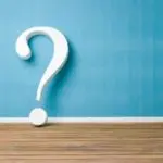 Large white question mark on a blue wall and wooden floor, representing plumbing inquiries by Allore's Plumbing Services.