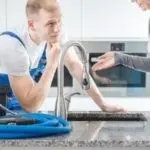 Technician in blue apron assisting customer with kitchen plumbing in a modern setting. Allore's Plumbing Services LLC.