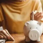 A person placing a coin in a friendly white piggy bank on a wooden table, promoting savings and financial awareness.