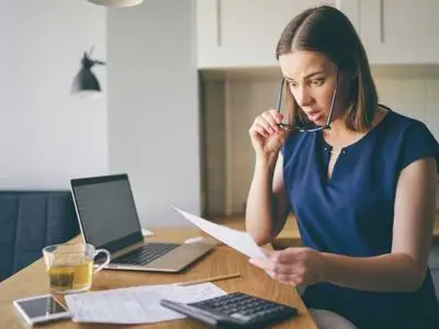 Woman reviewing water bill with concern at a desk, laptop and calculator present, highlighting issues related to fluctuating water expenses and potential plumbing leaks.