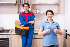 Licensed plumber in blue uniform with toolbox and satisfied homeowner giving thumbs up in modern kitchen, highlighting professional plumbing services and customer satisfaction.