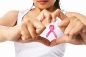 Woman forming a heart shape with hands, displaying a pink ribbon on her shirt, symbolizing breast cancer awareness and support.