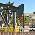 Oil pump jack in residential area of Stuart, FL, with safety fence and palm trees under a clear blue sky.