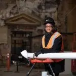 Person in safety gear reviewing white plans in construction environment with equipment and stone wall background.