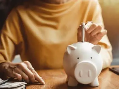 Person inserting a coin into a white piggy bank on a wooden table, symbolizing savings and financial planning.
