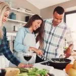 Three individuals cooking in a bright kitchen with utensils, fresh ingredients, a frying pan, and a glass of red wine.