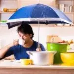Man at kitchen counter with blue umbrella, pots, and bowls showcasing plumbing tips and humor in home maintenance.