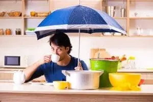 Man sitting at kitchen counter with an umbrella, drinking from a cup, surrounded by buckets and pots collecting water from a plumbing leak.
