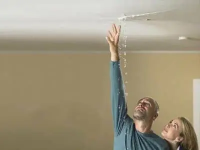 Man and woman looking up at ceiling leak, water dripping, indicating potential hidden plumbing issues affecting water pressure and household water bills.