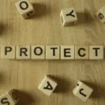 Scrabble-like tiles spelling 'PROTECT' on a wooden surface related to plumbing services by Allore's Plumbing.