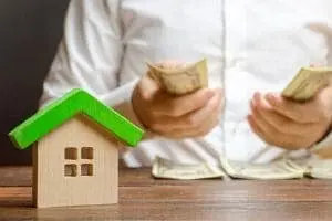 Person holding cash while counting bills, with a small wooden house model symbolizing home expenses and the financial impact of hard water on plumbing.
