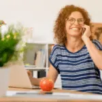 Woman with curly hair and glasses in home office on phone, representing Allore's Plumbing Services.