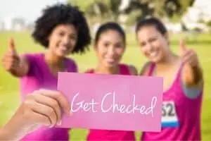 Three women in pink shirts smiling and giving thumbs up, holding a sign that says "Get Checked," promoting breast cancer awareness and the importance of early detection.