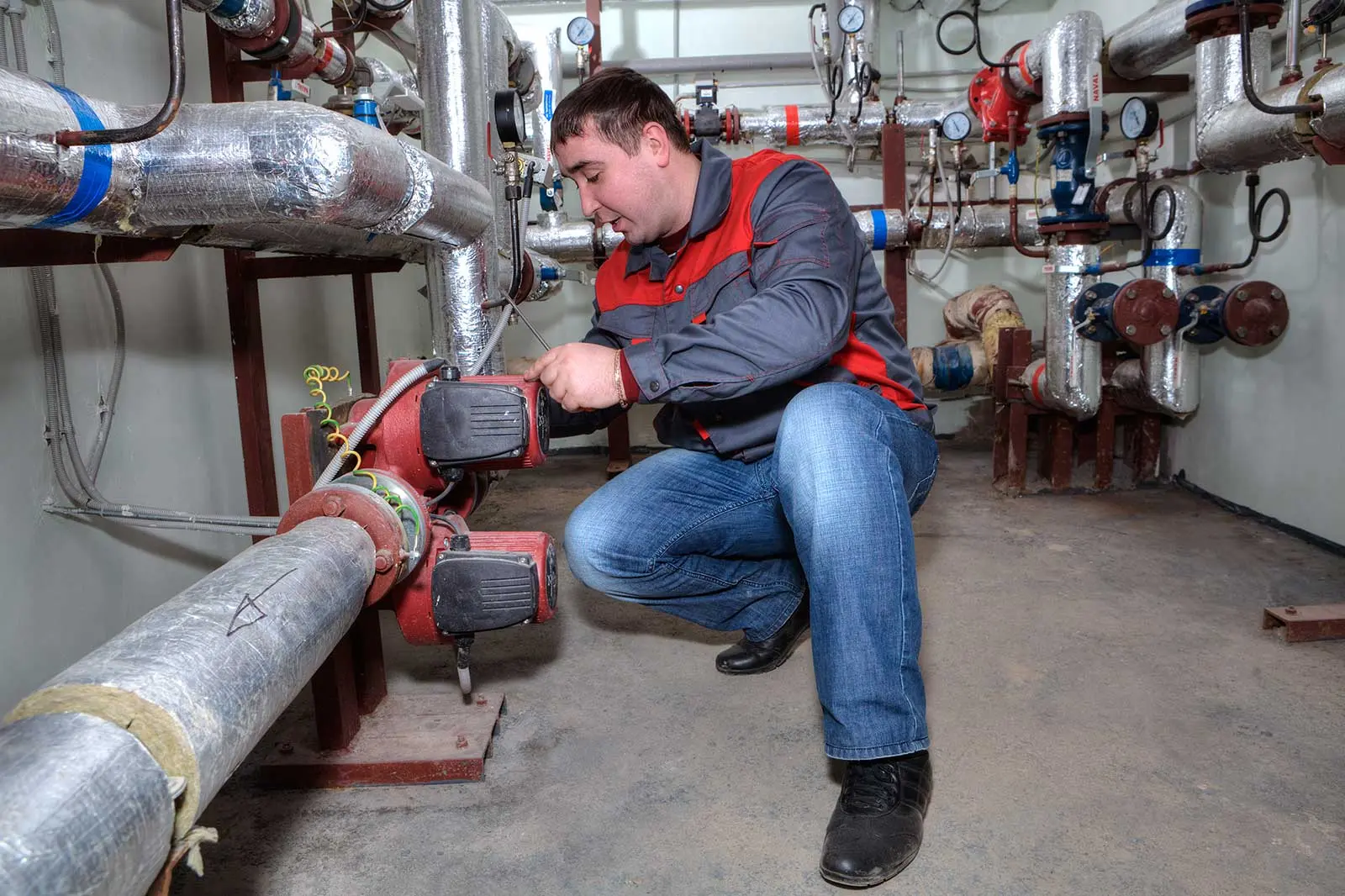 Technician repairing plumbing equipment by pipes and valves in a well-lit industrial setting for Allore's Plumbing Services.