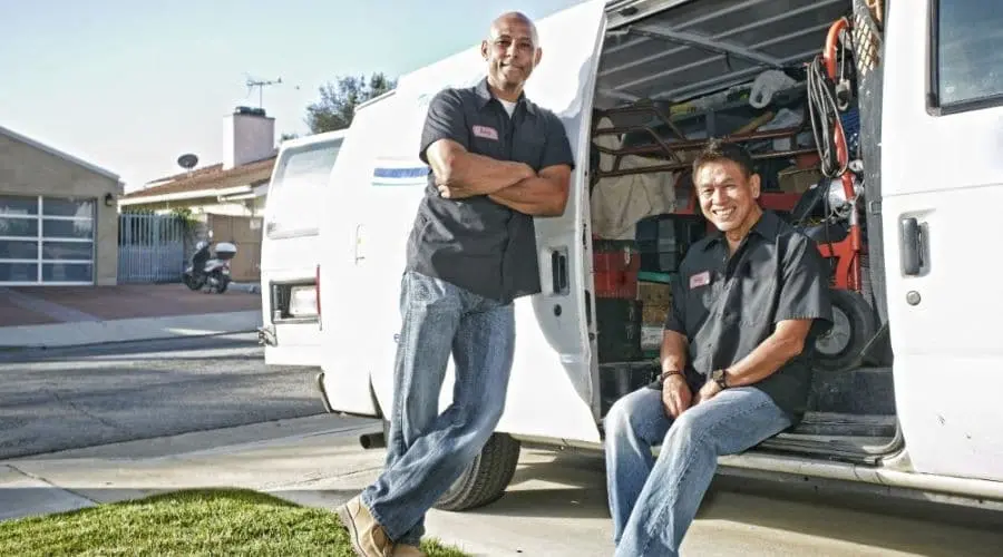 Two men in work uniforms by a service van, showcasing tools in a residential area under bright sunlight.