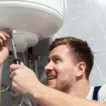 Smiling male plumber working under sink with adjustable wrench and under-sink water heater in modern bathroom.