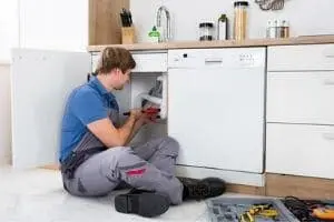 Plumber repairing kitchen sink undercounter, addressing faucet leak issues, with tools and plumbing supplies visible.