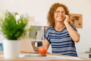 Woman talking on the phone at a desk with a laptop and an apple, representing the importance of communication and maintenance in plumbing services.