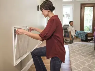 Woman replacing air filter in HVAC system, demonstrating routine maintenance for improved indoor air quality and system efficiency, with child playing in the background.