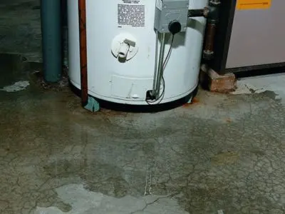 Leaking water heater with visible water stains on the floor, indicating potential plumbing issues and the need for replacement services.