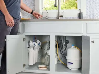 Person filling a glass with water from a kitchen sink, showcasing an under-sink reverse osmosis water filtration system, emphasizing home water quality improvement.