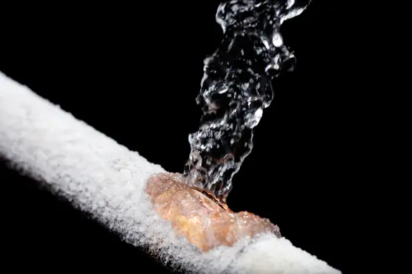 Frozen pipe with water leaking from a crack, surrounded by ice and frost, illustrating plumbing issues related to winter conditions.
