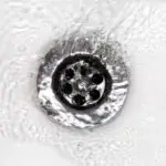 Circular metallic drain cover with holes, surrounded by flowing water, showcasing plumbing maintenance by A & W Plumbing.