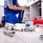 A plumber in blue overalls works on a kitchen cabinet with tools for A & W Plumbing and Heating, Inc.