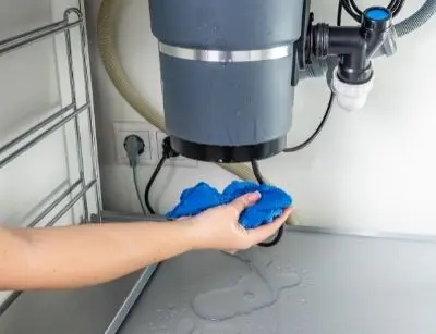 Hand holding a blue cloth near a leaking garbage disposal under a kitchen sink, illustrating signs of malfunction and the need for repair services.
