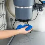 A hand holding a blue cleaning cloth near a sink with garbage disposal, showing plumbing maintenance for A & W Plumbing.