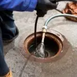 Plumber in work attire using a tool to spray water from a manhole opening, addressing outdoor plumbing issues during spring.
