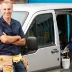 Smiling plumber with tool belt by van, showcasing tools for outdoor plumbing services by A & W Plumbing and Heating.