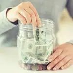 A person inserting cash and coins into a jar, representing savings for heating costs with B & D Plumbing, Heating & A/C.