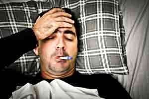 Man lying in bed with a thermometer in his mouth, appearing unwell and fatigued, emphasizing the importance of home health measures to combat flu spread.