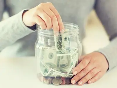Person placing cash into a jar filled with coins, symbolizing savings and financial management related to energy-efficient heating solutions.