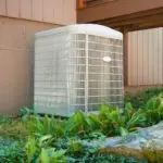 Air conditioning unit beside a building, surrounded by green foliage and a stone pathway, by B & D Plumbing, Heating & A/C.