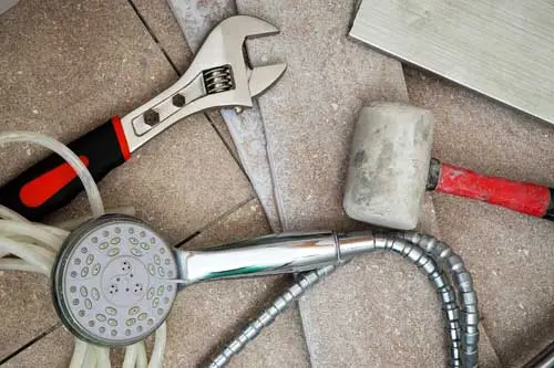 Plumbing tools including a wrench, rubber mallet, and showerhead on tiled surface, illustrating bathroom remodeling essentials.