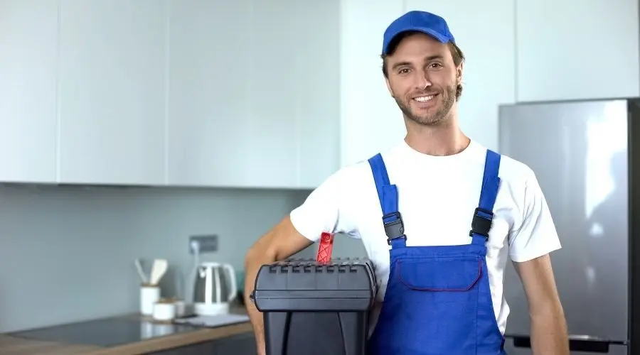 Young plumber in overalls and cap, smiling beside a toolbox in a modern kitchen setting of B & D Plumbing, Heating & A/C.