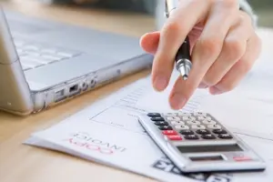 Person calculating costs with a calculator and laptop, reviewing air conditioning installation invoice for older homes.