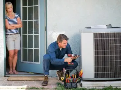HVAC technician repairing an air conditioning unit outside a home, with a homeowner observing, emphasizing the importance of professional air conditioner maintenance and repair services.