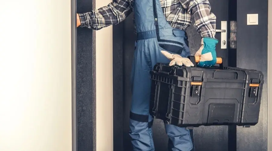 HVAC technician entering doorway with a black toolbox in plaid shirt and blue overalls for air conditioning maintenance.