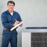 HVAC technician in blue outfit holding clipboard by air conditioning unit outdoors for Boutwell's Air Masters.