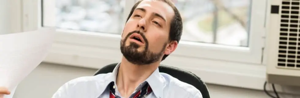 Man in a business shirt appearing overheated and fatigued in an office setting, with an air conditioner visible in the background, illustrating the need for effective cooling solutions during summer.