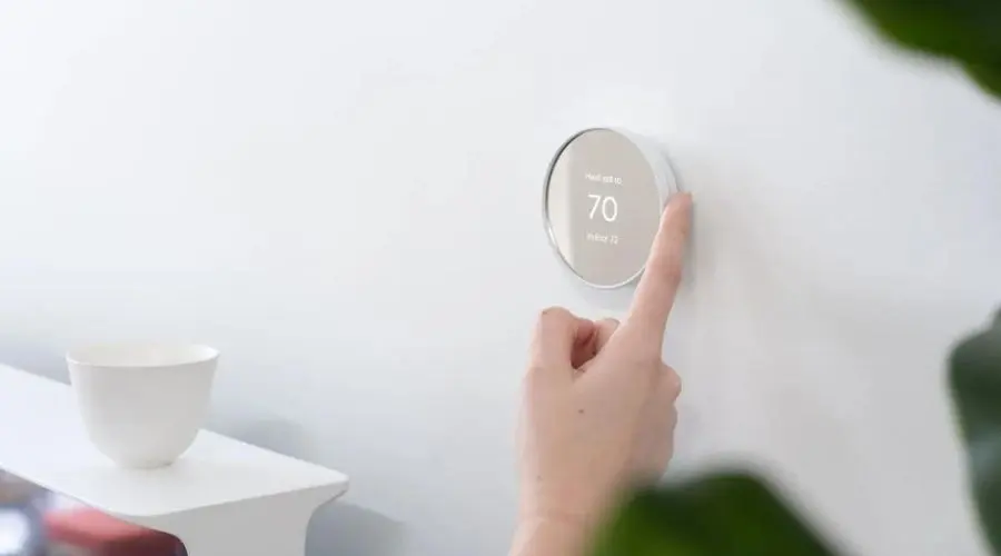 Smart thermostat being adjusted by a finger, showing 70 on a white wall, complemented by a green plant and simple bowl.