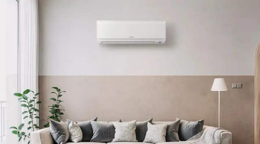 Wall-mounted air conditioner above a neutral-colored sofa with cushions, floor lamp, and potted plant for indoor comfort.