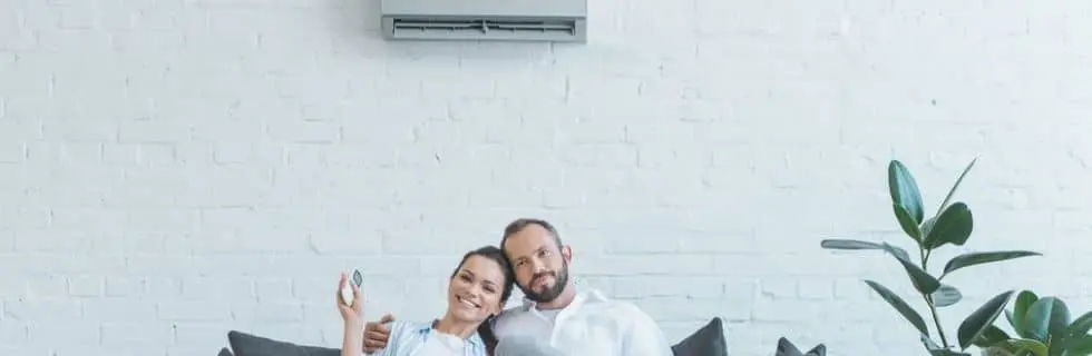 Couple relaxing on a couch with a remote control, enjoying cool air from a wall-mounted air conditioner in a modern indoor setting.