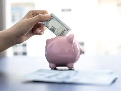 Hand inserting a hundred-dollar bill into a pink piggy bank, symbolizing savings and financial planning.