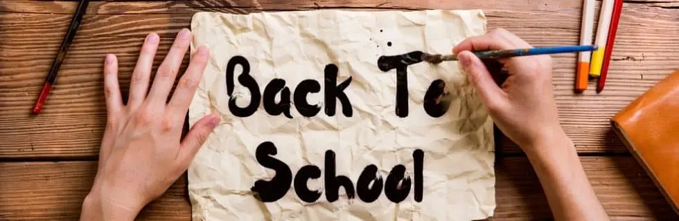 Hands painting "Back To School" on crumpled paper, surrounded by colorful markers and a notebook, symbolizing preparation for the upcoming school season.