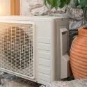 Air conditioning unit beside a decorative clay pot against a stone wall with plants, showcasing A-Plus HVAC solutions.