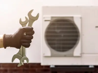 Hand holding wrenches in front of a heat pump unit, symbolizing heat pump repair and maintenance services.